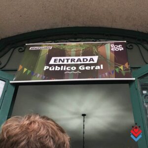 fotografia com foco no banner logo na entrada da feira! É um banner com o texto Entrada Público Geral. De fundo uma ilustração de um ambiente iluminado com luz verde, o pôster da feira.