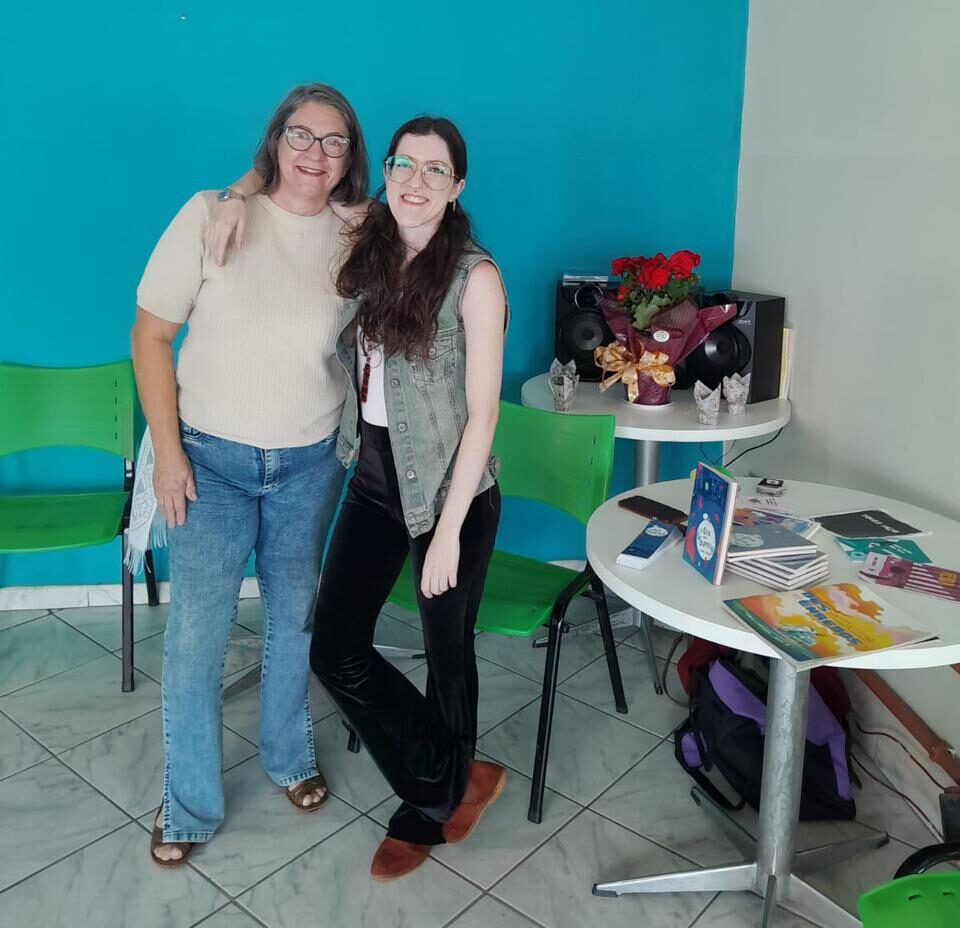 mesmo canto do salão da biblioteca, eu ao lado de minha mãe ao lado da mesa com meus livros. estou com meu braço por cima dela, abraçando, ambas sorrindo pra foto. 