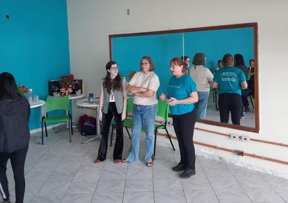 foto tirada mais de longe do salão, um espelho grande na parede atrás de 3 pessoas, eu, minha mãe e a bibliotecária rose. Rose diz algo enquanto nós duas prestamos atenção. Pelo espelho é possível ver que na nossa frente tem cadeiras e crianças sentadas. 